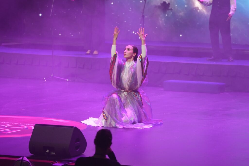 Dancer in a translucent costume kneels with arms raised under violet stage light