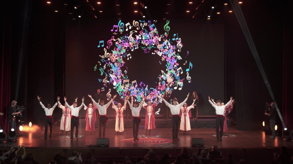Folk dancers raise their arms under a ring of colorful music notes on the LED screen