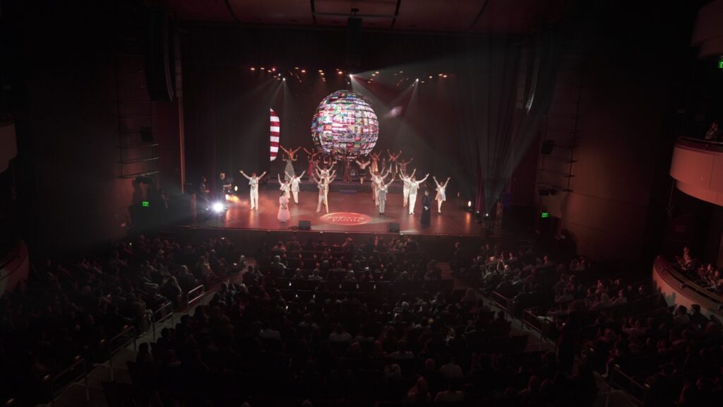 Wide view of Byers Theatre at Sandy Springs PAC as performers open Symphony of the Earth beneath a globe of world flags