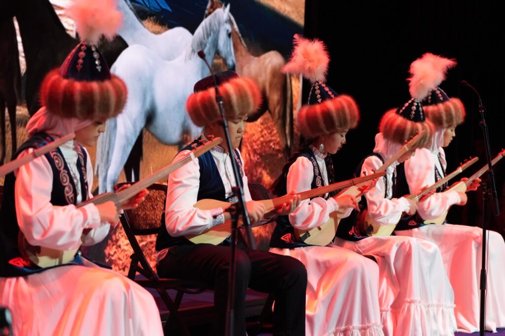 Young musicians in traditional fur hats play the komuz in front of a horse scene on the LED wall