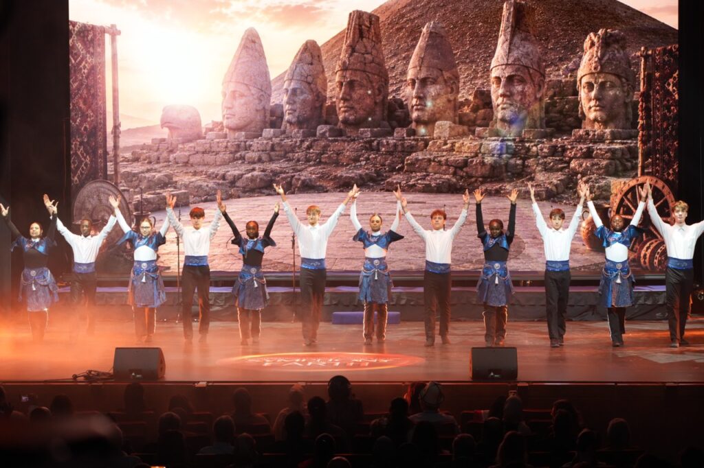 Dancers in blue and white costumes raise their arms in front of the Mount Nemrut statues on the LED screen