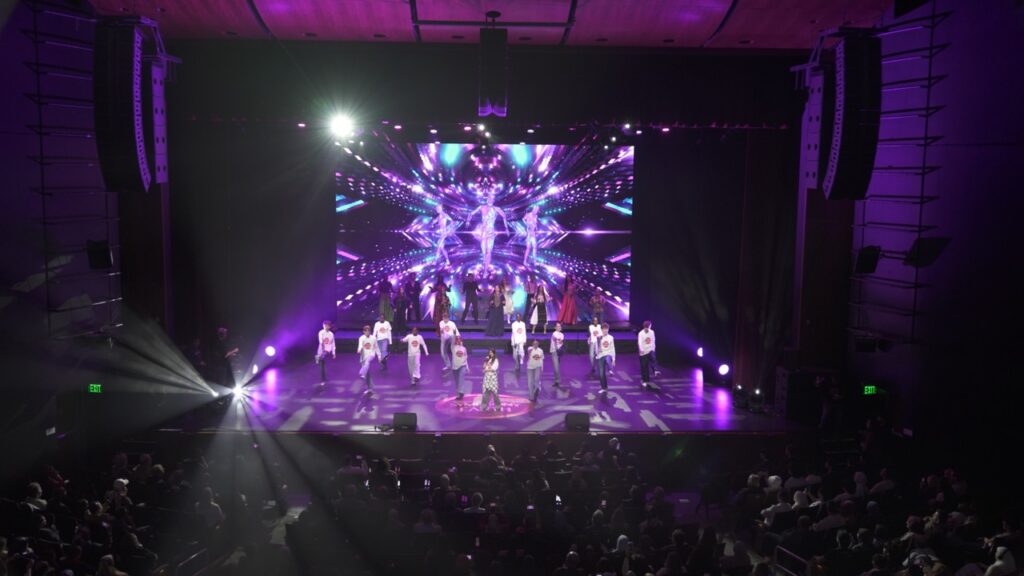 Performers in white shirts on a purple-lit stage with a mirrored LED backdrop at Byers Theatre