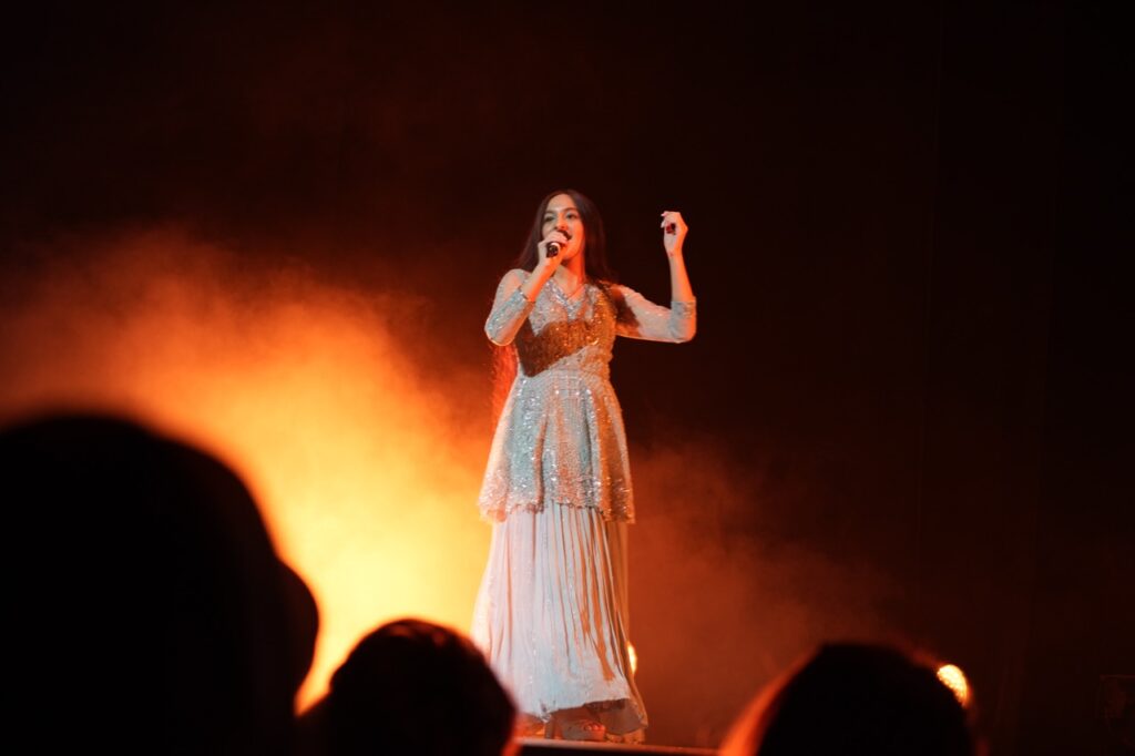 Singer in a sequined outfit performs through orange stage haze, audience silhouettes in the foreground