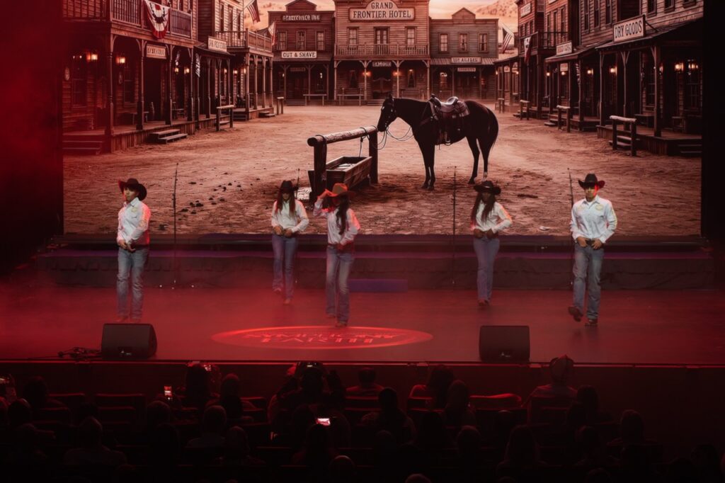 Performers in cowboy hats on stage with a Western frontier town on the LED screen